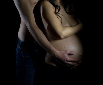 Midsection of shirtless man with hands on stomach of pregnant woman against black background