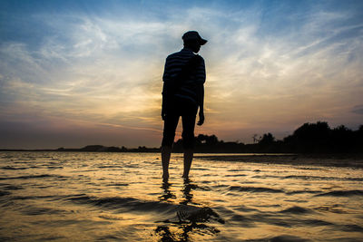 Rear view of man standing on shore against sunset sky