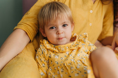 Little drooling girl in a yellow dress, leaning on mother, lies on the bed and looks at the camera