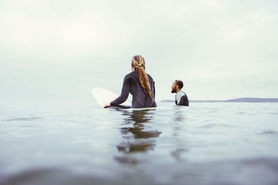 Male and female surfers relaxing in sea against cloudy sky