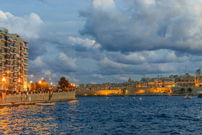 Illuminated buildings by sea against cloudy sky