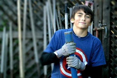 Portrait of young man holding metal rod while standing against fence