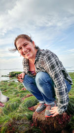 Portrait of smiling woman crouching on rock at beach against sky