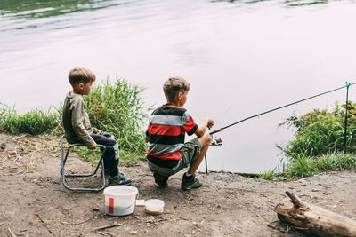 Two brothers sit on the shore of a lake or river and fish with fishing rods. 