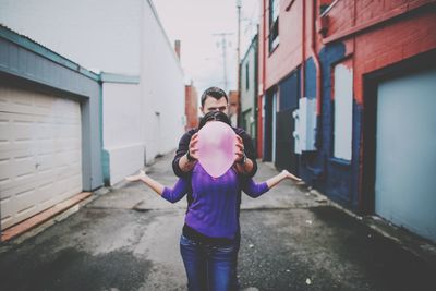 Man holding balloon over woman face while standing in alley