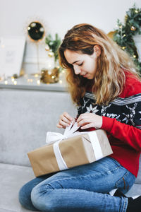 Pretty young girl opening presents near christmas tree. girl in red knitted sweater holds gift box.
