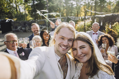 Portrait of happy newlywed couple taking selfie with guests in background on wedding day