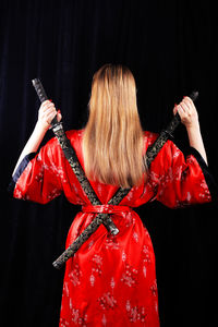 Rear view of woman wearing kimono and holding swords against black background