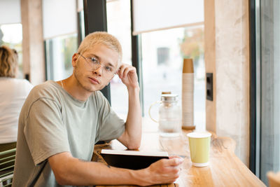 Stylish blond teenage boy 18-19 year old wear glasses reading paper book in cafe with coffee in cup.
