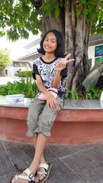 Portrait of happy girl showing peace sign against tree