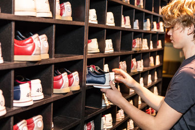 Side view of blond teenage boy holding bowling shoes at wooden rack