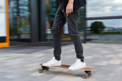 Shot of young man in black jeans and white sneakers riding a longboard in urban area.