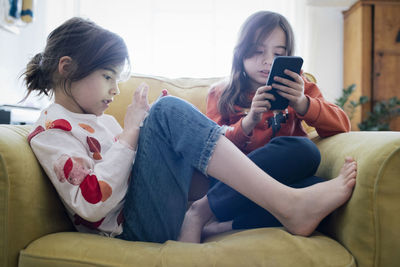 Girl using mobile phone sitting with sister on armchair at home