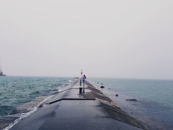 Distant view of teenage girl walking on pier in sea against clear sky