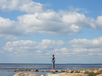 Beautiful girl standing on rocks by sea against sky