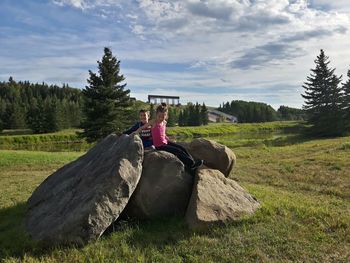 Siblings sitting on rocks at field against sky