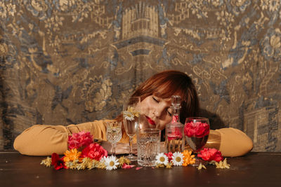 Young sad redhead female holding wineglass while sitting near table with floral and glassware arrangement with eyes closed