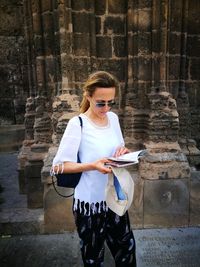 Young woman holding camera while standing against brick wall