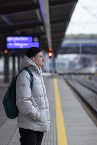 Side view of young man in  jacket with backpack standing and waiting for train at railway station.