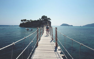 Rear view of man walking on footbridge over sea against clear sky