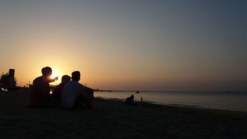 Silhouette people sitting on beach against sky during sunset