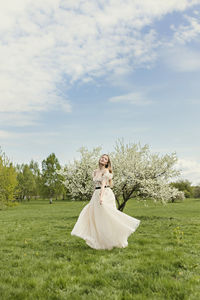 A young blonde in a long white dress poses near a cherry blossom in the garden, a spring landscape.