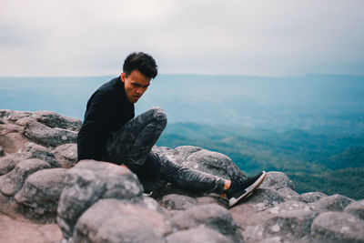 Young man sitting on rock by sea against sky