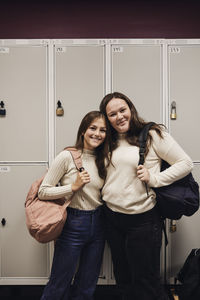 Portrait of smiling female students with backpacks standing against locker in school