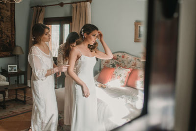 Attractive happy ladies helping young female to get into wedding dress near bed in bedroom