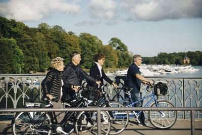 Senior male and female friends with bicycles walking on bridge over river