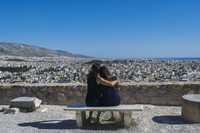 Rear view of two female friends sitting on bench against blue sky