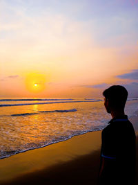 Side view of young man standing at beach against sky during sunset