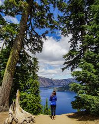 Rear view of woman standing on field by trees at crater lake national park against cloudy sky