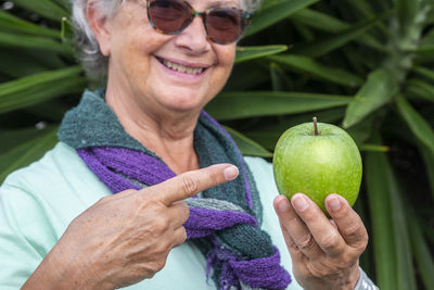 Smiling senior woman holding apple while standing against plant