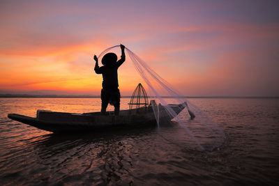 Silhouette fisherman holding fishing net in boat on sea during sunset