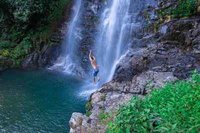 Young man jumping from cliff at natural waterfall blue water at morning from top angle