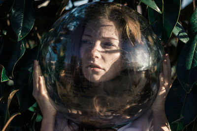 Close-up of young woman wearing glass ball on head against leaves