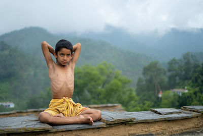 Portrait of shirtless man sitting on wood against mountain