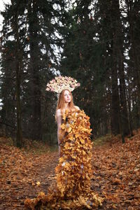 Thoughtful young woman covered with leaves standing at forest during autumn