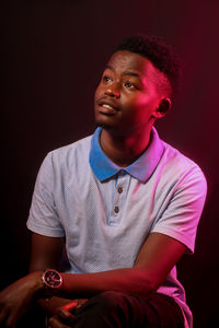 Young man looking away while sitting against black background