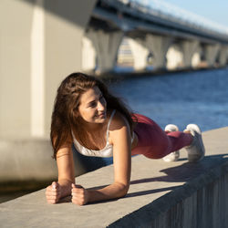 Full length of beautiful young woman exercising by sea against bridge