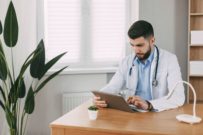A male doctor of oriental appearance is concentratedly working on a tablet, sitting at a table 