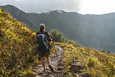 Rear view of young woman with big backpack trekking poles walking along mountain trail hiking