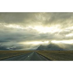 Road passing through field against cloudy sky