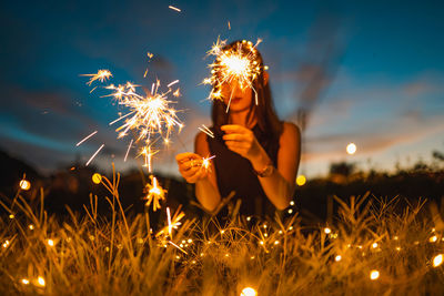 Woman with illuminated sparklers sitting on grass during sunset