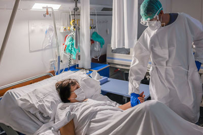Professional male doctor in medical uniform and gloves visiting and examining senior female patient lying on bed in protective mask in hospital ward while curing from coronavirus