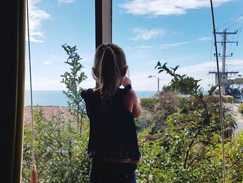 Rear view of woman standing by window against sky