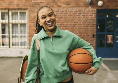 Portrait of happy young woman holding basketball under arm while standing in front of building