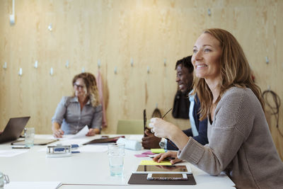 Smiling businesswoman looking away while sitting with colleagues at conference table in board room