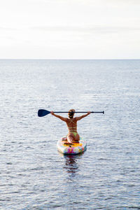 Active female surfer in swimwear holding paddle in raised hand while standing on paddle board floating in sea water near coast